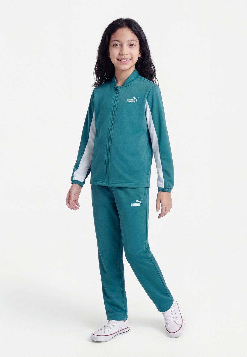 Junges Mädchen lächelnd und stehend, trägt einen türkisfarbenen Puma-Trainingsanzug mit weißen Streifen und weiße Turnschuhe vor weißem Hintergrund.