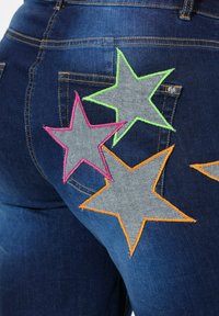 Jeans de mezclilla azul con grandes aplicaciones en forma de estrella en verde, rosa y naranja, con costuras en contraste, situadas en los bolsillos traseros.