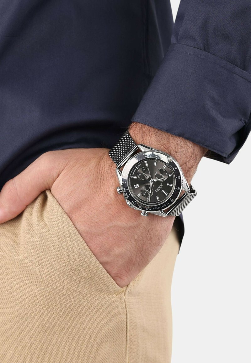 Männliche Hand in einer beigen Hosentasche, die eine silberne Metall-Mesh-Armbanduhr mit schwarzem Zifferblatt und Chronographen-Details trägt, während einen navyblauen Hemdsärmel sichtbar ist.