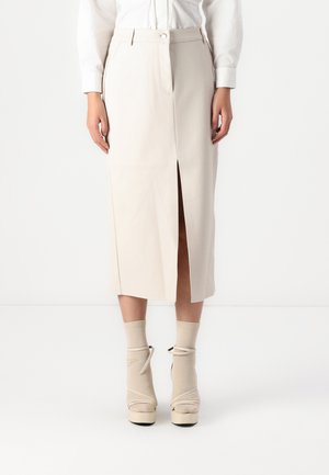 Kobieta ubrana w beżową midi spódnicę z wysokim stanem i rozcięciem z przodu, białą koszulę z długim rękawem włożoną do środka, beżowe skarpetki i beżowe buty na platformie.