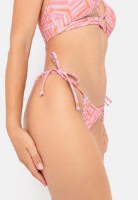 Bikini komplekts ar rozā ģeometrisku rakstu. Augšdaļa ir mezglota dizaina; apakšdaļa sasaistās sānos ar auklām. Gluda auduma tekstūra.