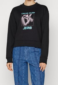 Černý zkrácený svetr s kulatým výstřihem, zdobený multibarevným grafickým prvkem "CK" a "JEANS." V kombinaci s modrými vzorovanými kalhotami.