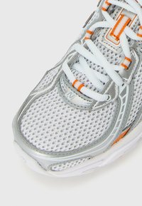 Chaussure de sport blanche et argentée avec une tige en maille et des lacets blancs, présentant des accents orange.