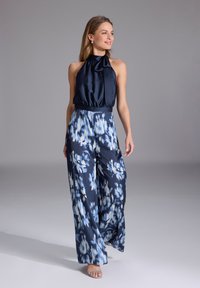 Mono de halter azul marino con cuello atado, que presenta pantalones de pierna ancha con un estampado en azul y blanco. Acabado de satén suave, diseño casual pero elegante.