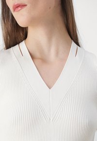 Maglione bianco a coste con scollatura a V profonda e fessure sulle spalle, caratterizzato da un motivo testurizzato e una vestibilità aderente.