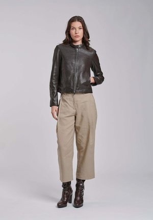 Kobieta stoi, ubrana w czarną skórzaną kurtkę z fakturą, beżowe szerokie spodnie i ciemnobrązowe botki na obcasie na jednolitym jasnym tle.