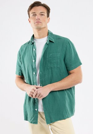 Homme portant une chemise verte à manches courtes déboutonnée par-dessus un t-shirt blanc et un pantalon beige, debout les mains jointes devant un fond blanc.