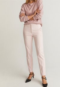 Světle růžové přiléhavé kalhoty s hladkou texturou a bočními kapsami, kombinované se světle růžovým svetrem a černými lodičkami s špičatou špičkou.