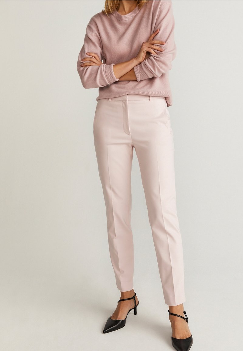 Světle růžové přiléhavé kalhoty s hladkou texturou a bočními kapsami, kombinované se světle růžovým svetrem a černými lodičkami s špičatou špičkou.