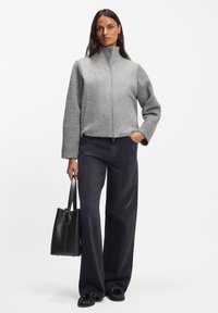 Hall triiksärgi stiilis jope tekstuursete varrukatega, ühendatud mustade laiade pükstega. Modell hoiab musta käekotti, seistes valge tausta ees.