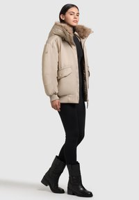 Beige vadderad jacka med en huva klädd i päls, dragkedjestängning och sidofickor, ihop med svarta leggings och knähöga svarta läderstövlar.