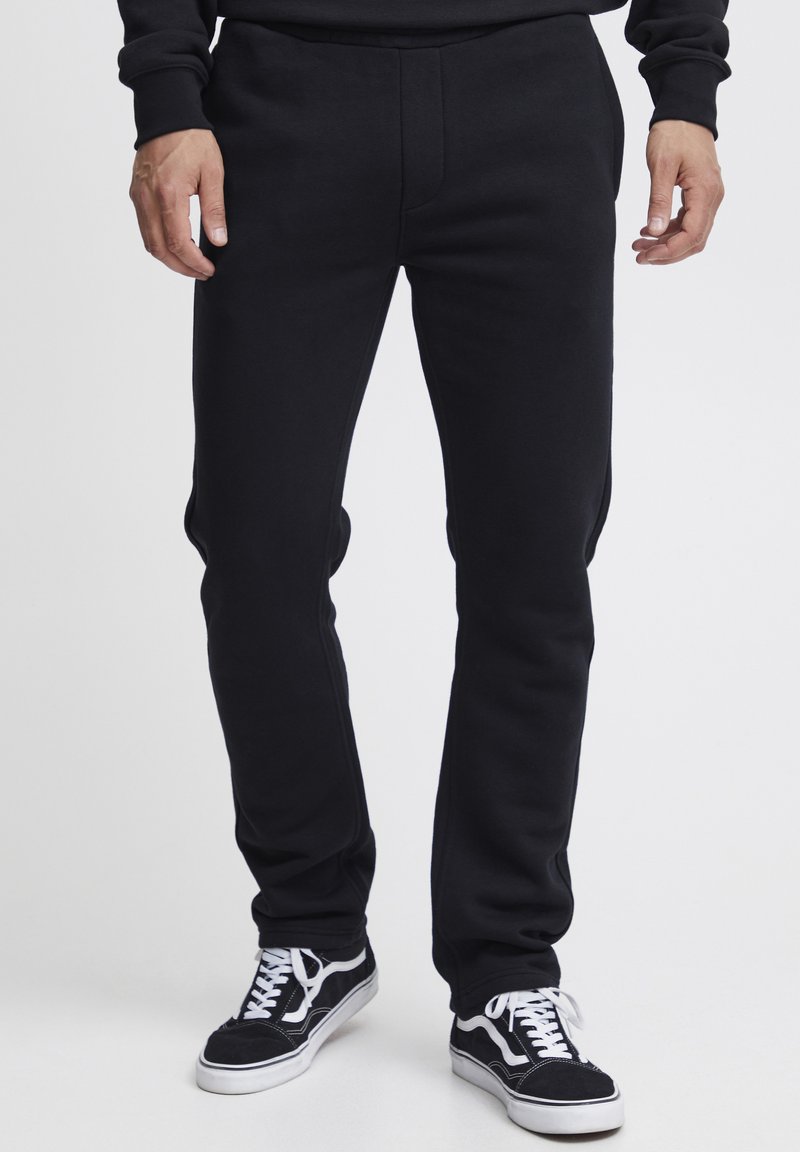 Black sweatpants in a slim fit, featuring a smooth texture, stitched detailing, and an elastic waistband, paired with black and white sneakers.