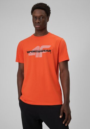 Mężczyzna stojący, ubrany w pomarańczowy t-shirt z krótkim rękawem z logo "4F SPORTSWEAR" i czarne spodnie na jednolitym szarym tle.