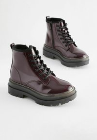 Bottines en cuir verni bordeaux avec lacets noirs, semelles en caoutchouc noir épaisses, fermeture éclair sur le côté et col rembourré pour le confort.
