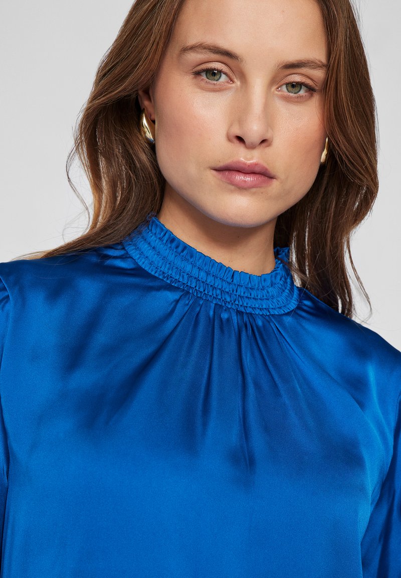 Blauwe satijnen blouse met een hoge geplooide kraag en gerimpelde details. Gladde textuur, lange mouwen, met een zachte, vloeiende val.