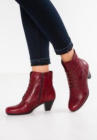 Botas de tobillo de cuero rojo con punta redondeada, cremallera lateral y detalle de cordones; tacón modesto; acabado texturizado; combinadas con jeans de mezclilla oscura.