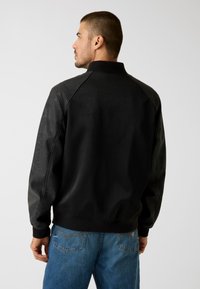Zwarte bomberjack met een gladde voorkant, leren mouwen en geribbelde boorden. De achterkant heeft een strak ontwerp zonder zichtbare logo's of patronen.