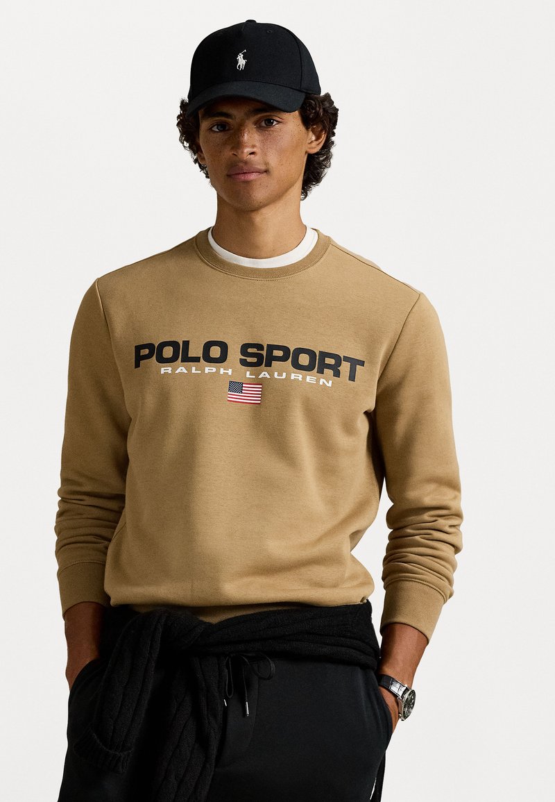 Brūns džemperis ar uzrakstu "POLO SPORT" un American flag grafiku, apvienots ar melnu cepuri un melnām biksēm, kurām ir adīts jostasvieta.