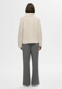 Bež velik pulover s turtleneck iz teksturiranega pletenega vzorca, v kombinaciji s sivimi širokimi hlačami. Enostavna ozadje.