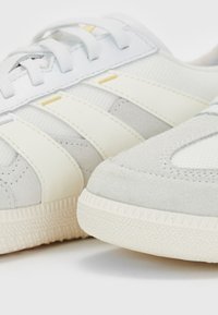 Baskets blanches et gris clair en daim et mailles, avec trois rayures en crème et une semelle en caoutchouc texturée.