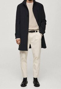 Cappotto di lana scuro, maglione beige a coste, pantaloni crema, cintura nera. Scarpe in pelle nera con superficie texturizzata. Vestibilità slim, silhouette moderna.