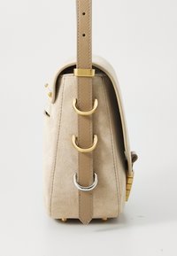 Borsa in suede beige vista di lato con tracolla in cuoio beige infilata attraverso tre anelli a D di metallo, due dorati e uno argentato, su sfondo bianco.