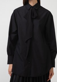 Blusa nera con colletto e un fiocco legato al collo. Maniche lunghe con polsi abbottonati, realizzata in tessuto liscio. Vestibilità classica e rilassata.