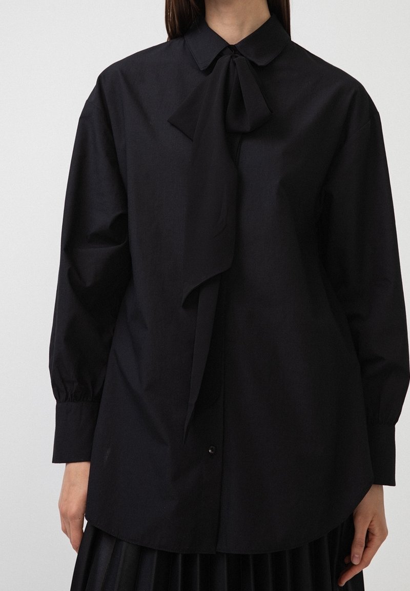 Blusa nera con colletto e un fiocco legato al collo. Maniche lunghe con polsi abbottonati, realizzata in tessuto liscio. Vestibilità classica e rilassata.