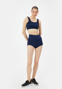 Sujetador deportivo de color azul marino y pantalones cortos de cintura alta, con textura acanalada y diseño ajustado. Llevado con zapatos deportivos negros, el modelo adopta una pose neutral.