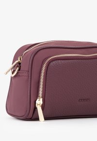 Borsa pochette in pelle bordeaux con zip dorate, forma rettangolare, superficie testurizzata e logo impresso sul lato.