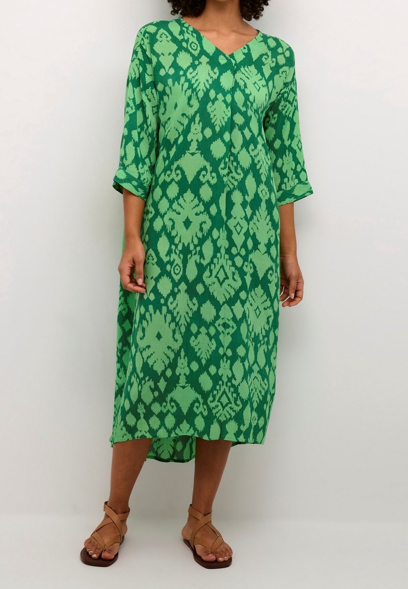 Femme portant une robe midi verte à motifs avec un décolleté en V et des manches trois-quarts, associée à des sandales plates à lanières beige.