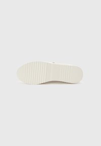Semelle de chaussure blanche avec un motif strié et un talon plat, photographiée sur un fond uni gris clair.