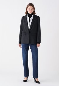 Blazer negro a rayas con solapa muesca, usado sobre una camisa blanca; combinado con jeans de mezclilla oscuros y zapatos de tacón negros.