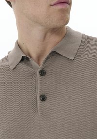 Camisa polo texturada en color beige con un patrón ondulado, dos botones negros en el cuello, mangas cortas y un diseño de cuello clásico.