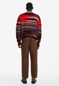 Strickpullover mit roten, blauen und braunen Streifen sowie Texturvariationen; kombiniert mit braunen weiten Hosen und braunen Schuhen.