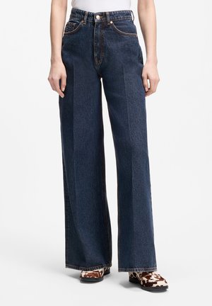 Femme portant un jean bleu foncé taille haute à jambes larges avec des coutures orange et des chaussures à imprimé animalier sur fond blanc.