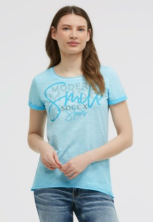 Junge Frau mit langen braunen Haaren, die ein hellblaues T-Shirt mit Grafik und blaue Jeans trägt, steht vor einem neutralen Hintergrund.