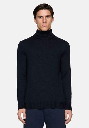 Uomo con capelli corti castani che indossa un maglione a collo alto nero a coste aderente e pantaloni scuri, in piedi contro uno sfondo chiaro e uniforme.