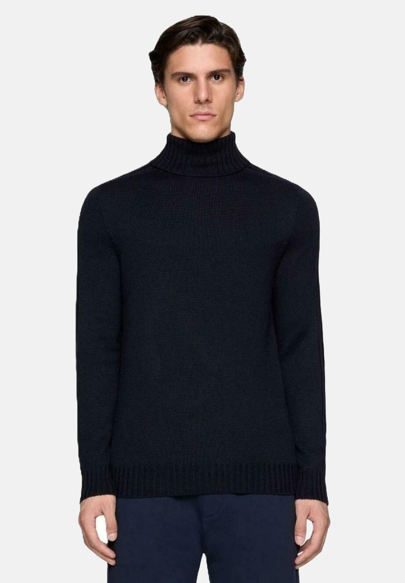 Uomo con capelli corti castani che indossa un maglione a collo alto nero a coste aderente e pantaloni scuri, in piedi contro uno sfondo chiaro e uniforme.