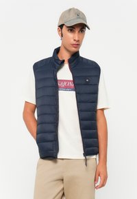 Marineblå dunvest med vertikal quiltet design, høy krage og trykknappdetaljer, kombinert med en lyse t-skjorte og beige bukser.