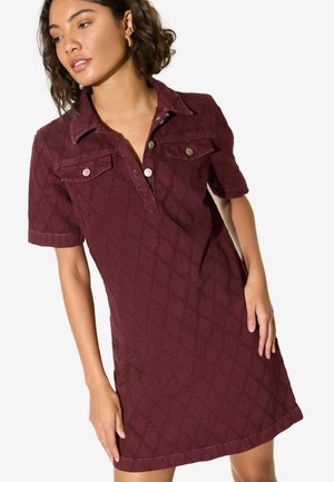 Mujer vestida con un vestido burdeos de manga corta y botones, con un sutil patrón de diamantes y bolsillos en el pecho, de pie frente a un fondo liso.