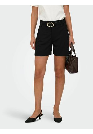 Femme portant un short noir ceinturé, un haut blanc, des talons slingback tissés noirs, tenant un sac à main marron foncé à ses côtés sur un fond uni.