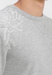 Felpa grigia in tessuto morbido con ricamo floreale bianco sulla spalla sinistra, scollo rotondo e polsini a costine.