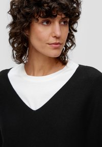Schwarzer Strickpullover mit V-Ausschnitt, über einem weißen Rundhals-Shirt getragen. Lockiges Haar umrahmt das Gesicht, ohne erkennbare Muster oder Akzente.