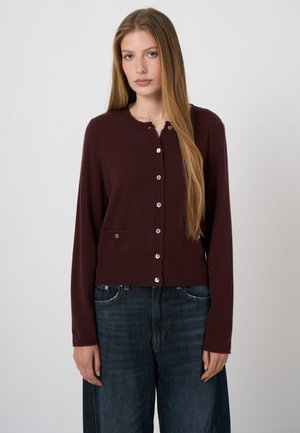 Jeune femme aux longs cheveux blonds portant un cardigan bordeaux foncé boutonné et un jean bleu foncé, debout devant un fond clair uni.