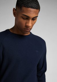 Pull en maille bleu marine avec un col rond et un logo brodé. Le tissu présente un motif texturé et des manches longues.