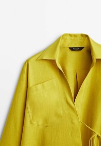 Valkoinen liinapaita kirkkaan keltaisena, jossa on terävä kaulus, yksi tasku ja sidontayksityiskohta. Paidassa on tekstuuripinta ja rento istuvuus.