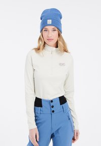 Crème kleurige lange mouwen zip-up top met een "Laten we gaan skiën" graphic, gecombineerd met hoge blauwe skibroeken met een zwarte tailleband en knopen.