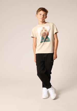 T-shirt beige avec un motif de chien, pantalon cargo noir et baskets blanches. Le t-shirt a des manches courtes et un col rond.