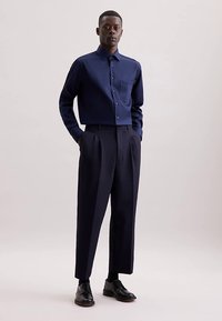 Camicia blu navy con bottoni e tasca, abbinata a pantaloni a vita alta e plissettati dello stesso colore blu navy. Scarpe in pelle nera completano il look.